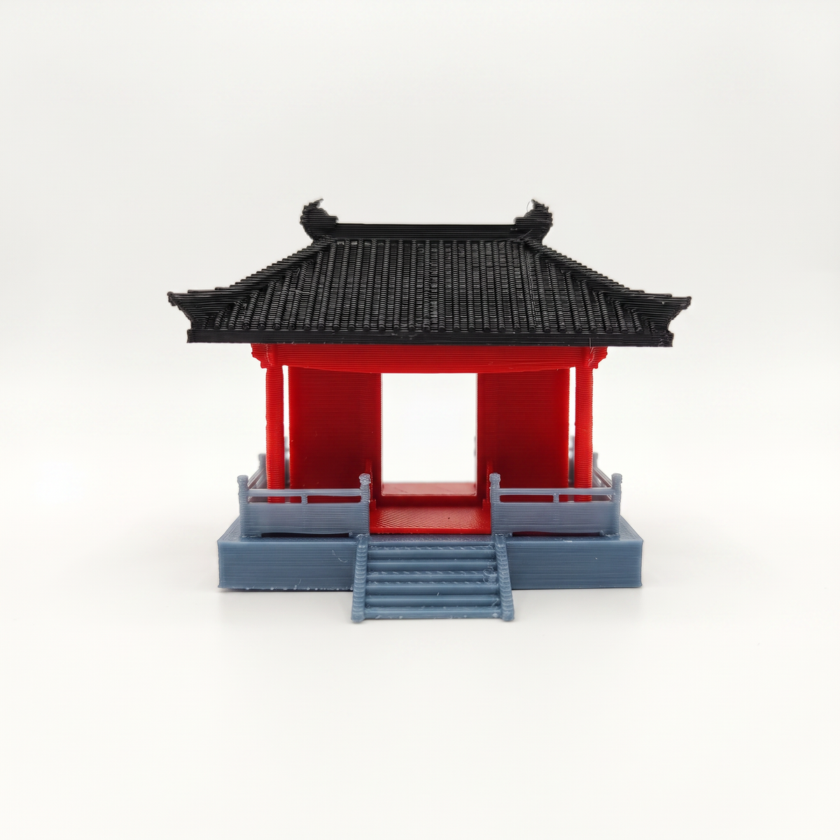 A miniature traditional East Asian pavilion model with a black tiled roof, red walls, and a blue base, shown on a seamless white background.
